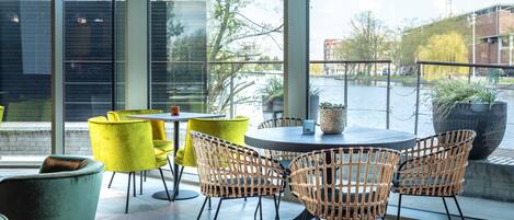 Un área de estar moderna al aire libre con mesas redondas y sillas de mimbre, amplias ventanas con vistas a un río y edificios.