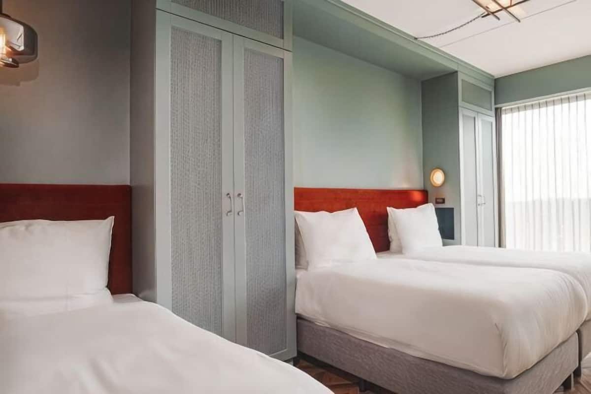 Habitación de hotel con dos camas, un armario, una mesita de noche con lámpara y una ventana con persianas.