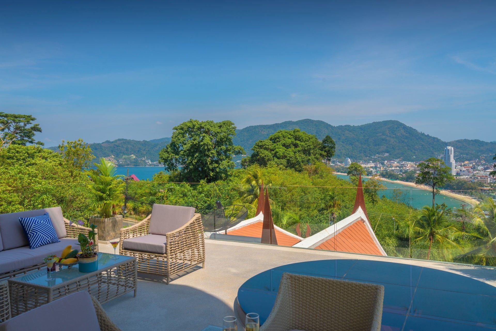 Une terrasse dotée de meubles en osier, d’une table en verre et offrant une vue sur une plage et des montagnes.
