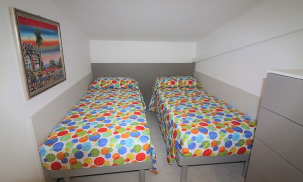 Una habitación pequeña y limpia con dos camas individuales, un cabecero gris y una pared blanca.