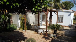 Een wit gebouw met een patio, inclusief een tafel en stoelen, omringd door verschillende planten en een pergola.
