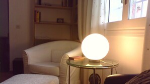 Een gezellige kamer met een tafellamp, een witte fauteuil, een klein rond tafeltje en een boekenkast.