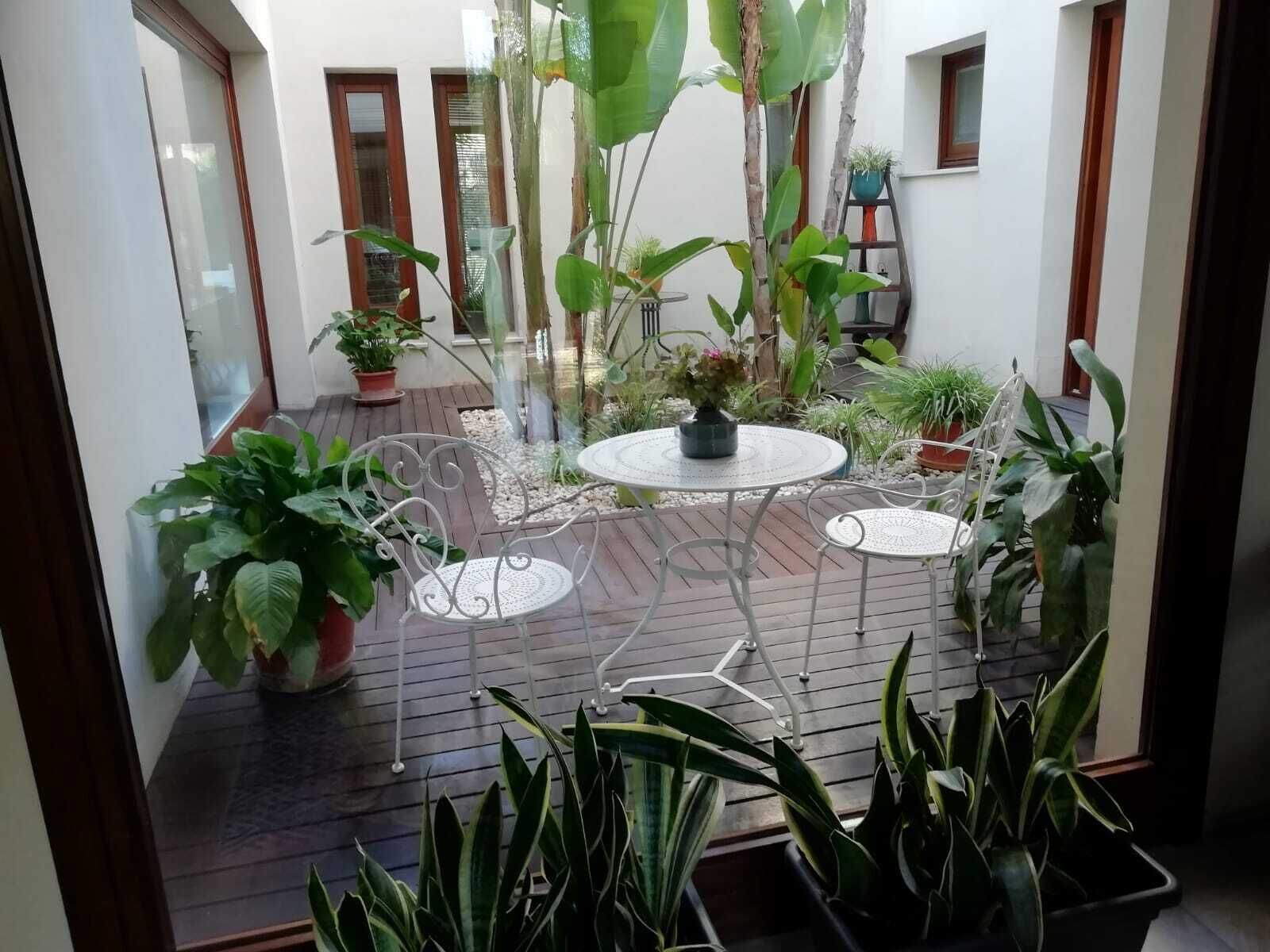 Un patio con una mesa redonda y sillas, rodeado de plantas en macetas y una terraza de madera.