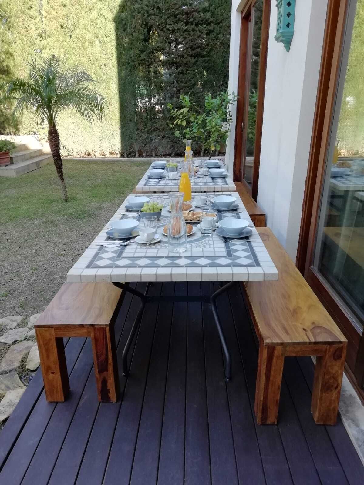 Une table de patio, prête pour le repas, avec un plateau carrelé, des bancs en bois et une carafe en verre remplie de jus d’orange.