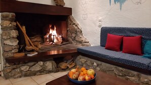 Un camino in pietra con un fuoco acceso, una parete in pietra con un affresco, un divano blu con motivi decorativi e cuscini rossi, e un tavolo con una ciotola di frutta.