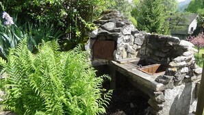 Un forno esterno in pietra con porta in legno, circondato da vegetazione lussureggiante e un muro in pietra.