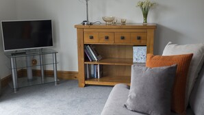 Sala de estar com um sofá cinza, uma mesa lateral de madeira com uma TV, livros e objetos decorativos.