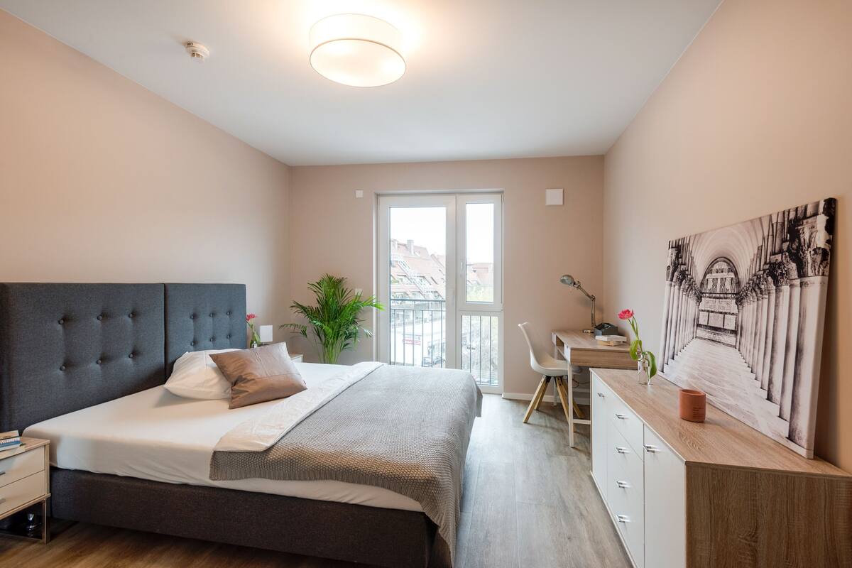 A bedroom with a bed, bedside table, desk, chair, and a large window.