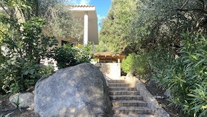 Un escalier en pierre mène à un bâtiment doté d’un toit en tuiles, qui est entouré de verdure et de gros rochers.