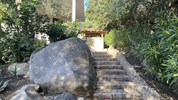 Un escalier en pierre mène à un bâtiment doté d’un toit en tuiles, qui est entouré de verdure et de gros rochers.