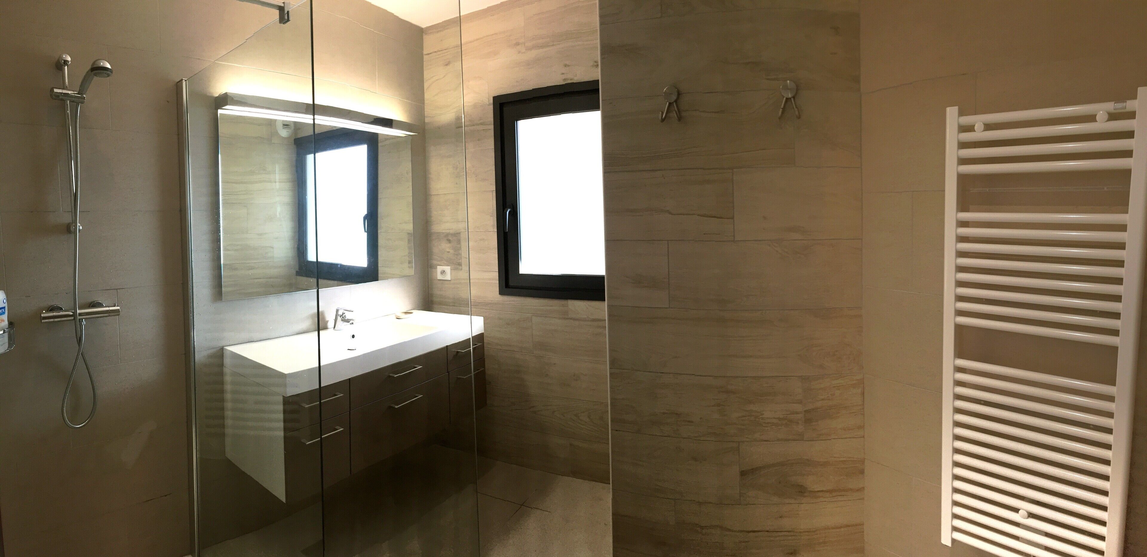 Een moderne badkamer met een douchecabine van glas, een grote spiegel, een wit wastafelmeubel en een ingebouwde verwarming.