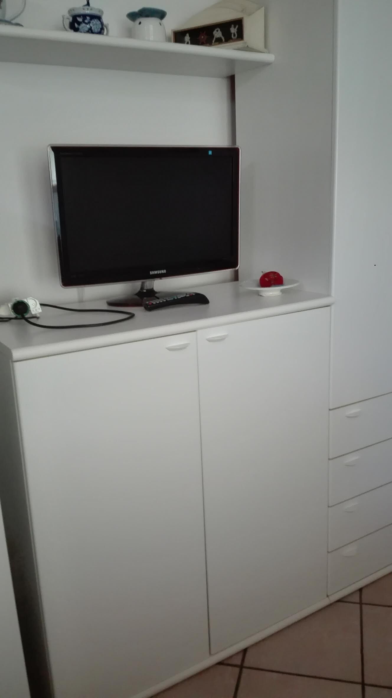A white kitchen with a Samsung TV mounted on the wall, a coffee maker, and a small shelf with decorative items.