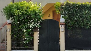Una casa de dos pisos con un portón negro, un balcón pequeño y un jardín con plantas blancas florecientes.