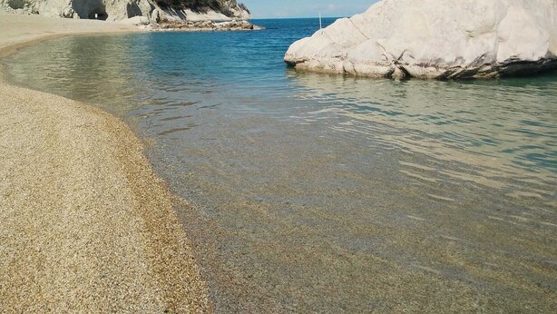 Una playa con aguas claras y poco profundas, un acantilado rocoso y una gran formación rocosa blanca.