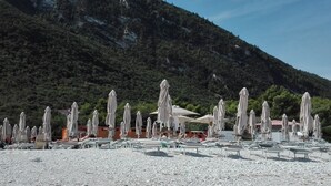 Una playa con numerosas tumbonas y sombrillas, arena de guijarros y montañas al fondo.