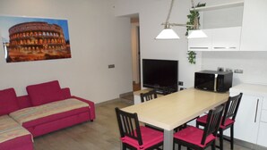 A modern living room with a sofa, a dining table with chairs, a flat-screen TV, and a kitchen area with cabinets and appliances.