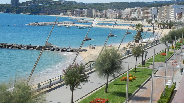 Un paseo marítimo frente a la playa, un rompeolas rocoso y una hilera de edificios al fondo.