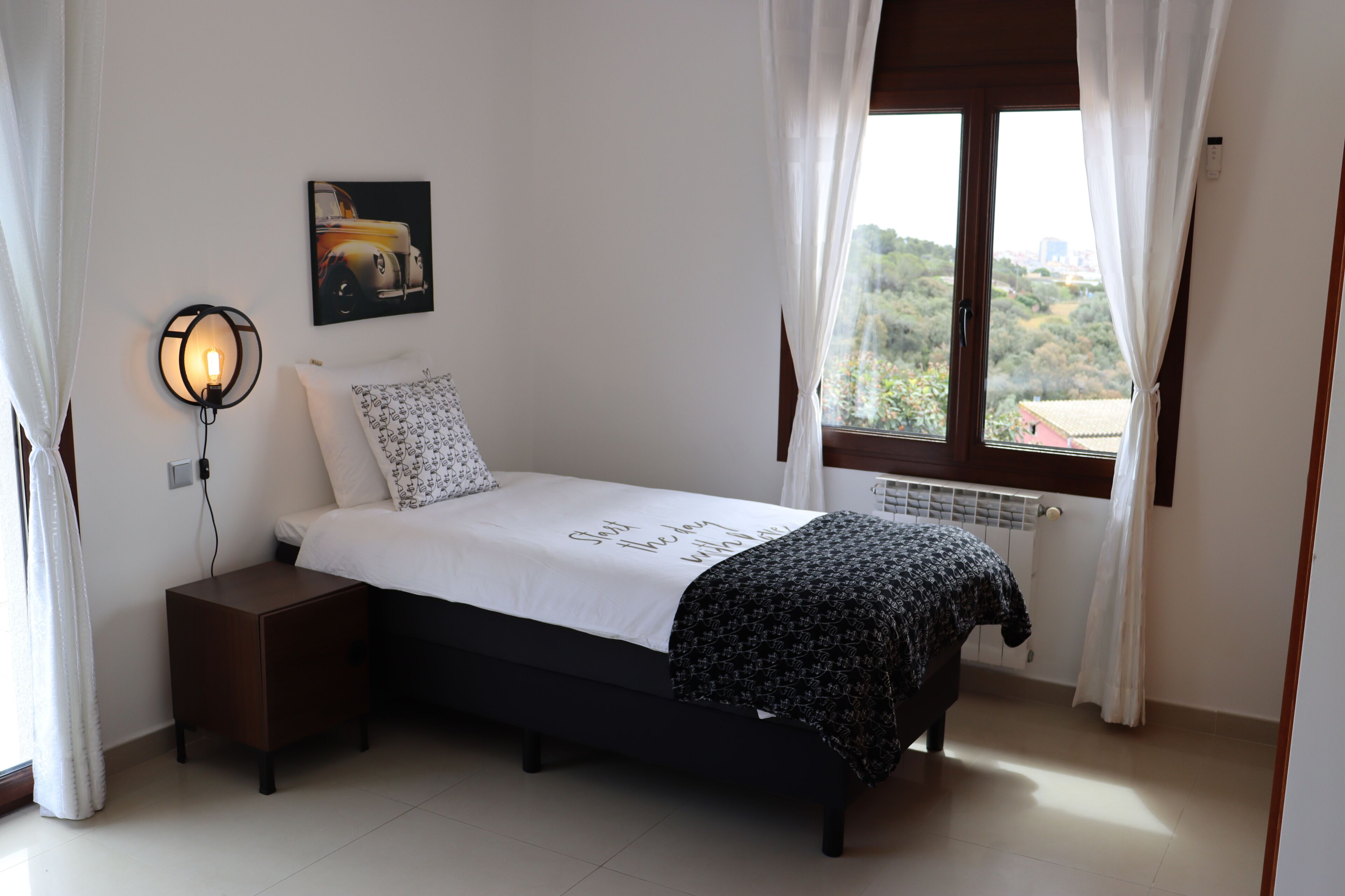 Une chambre à coucher comprenant un lit, une table de chevet, une lampe fixée au mur, une fenêtre avec des rideaux et une vue sur la verdure à l’extérieur.