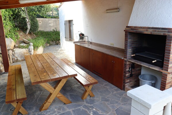 An outdoor patio with a wooden table and benches, a brick barbecue grill, and a stone floor. - Villa in beautiful location with sea views, heated pool, high-speed WIFI (Calonge)