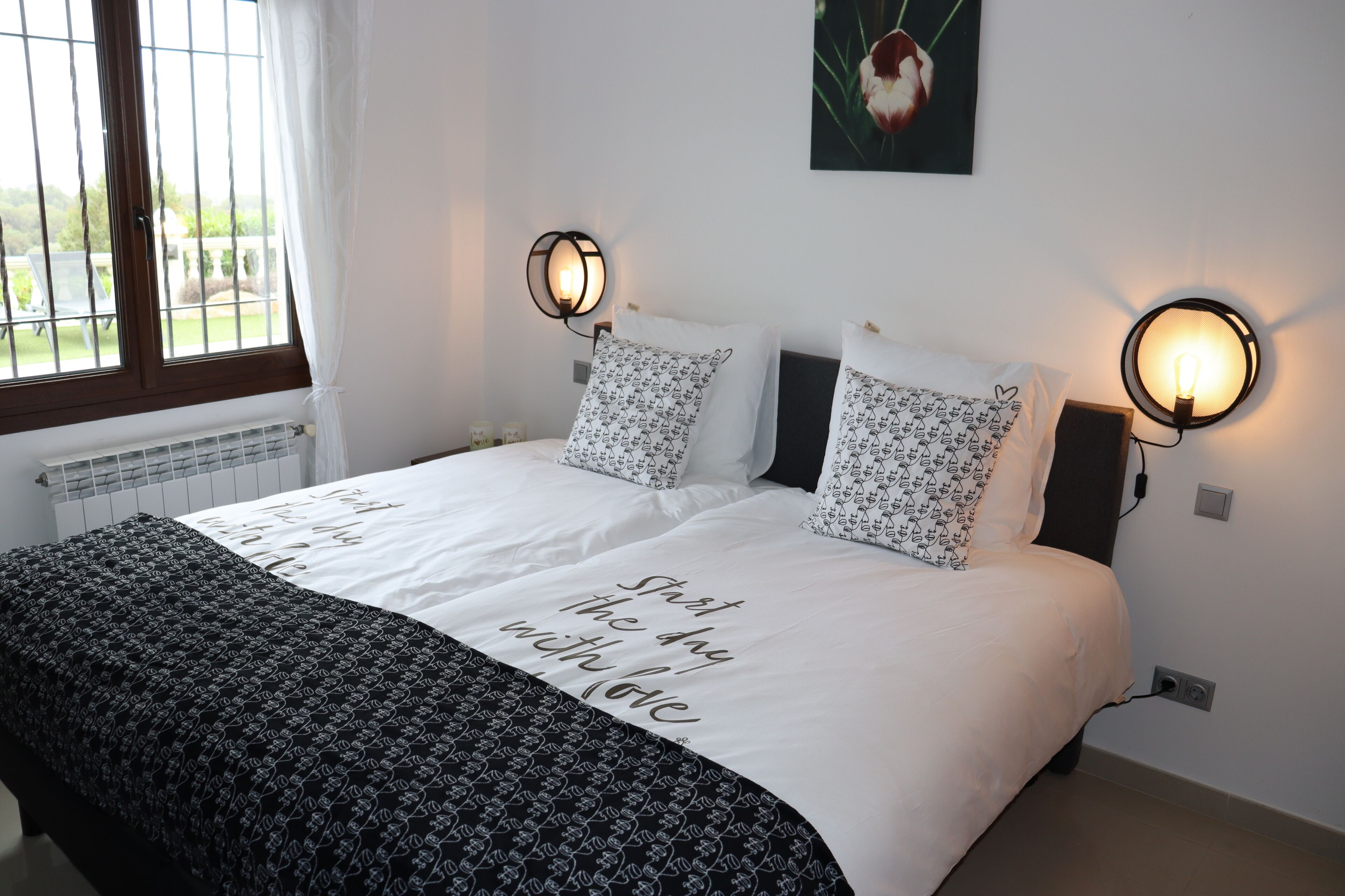 Un lit bien fait, avec une couette blanche et des coussins à motifs, deux lampes de lecture fixées au mur, un tableau encadré accroché au mur et une fenêtre avec des rideaux.
