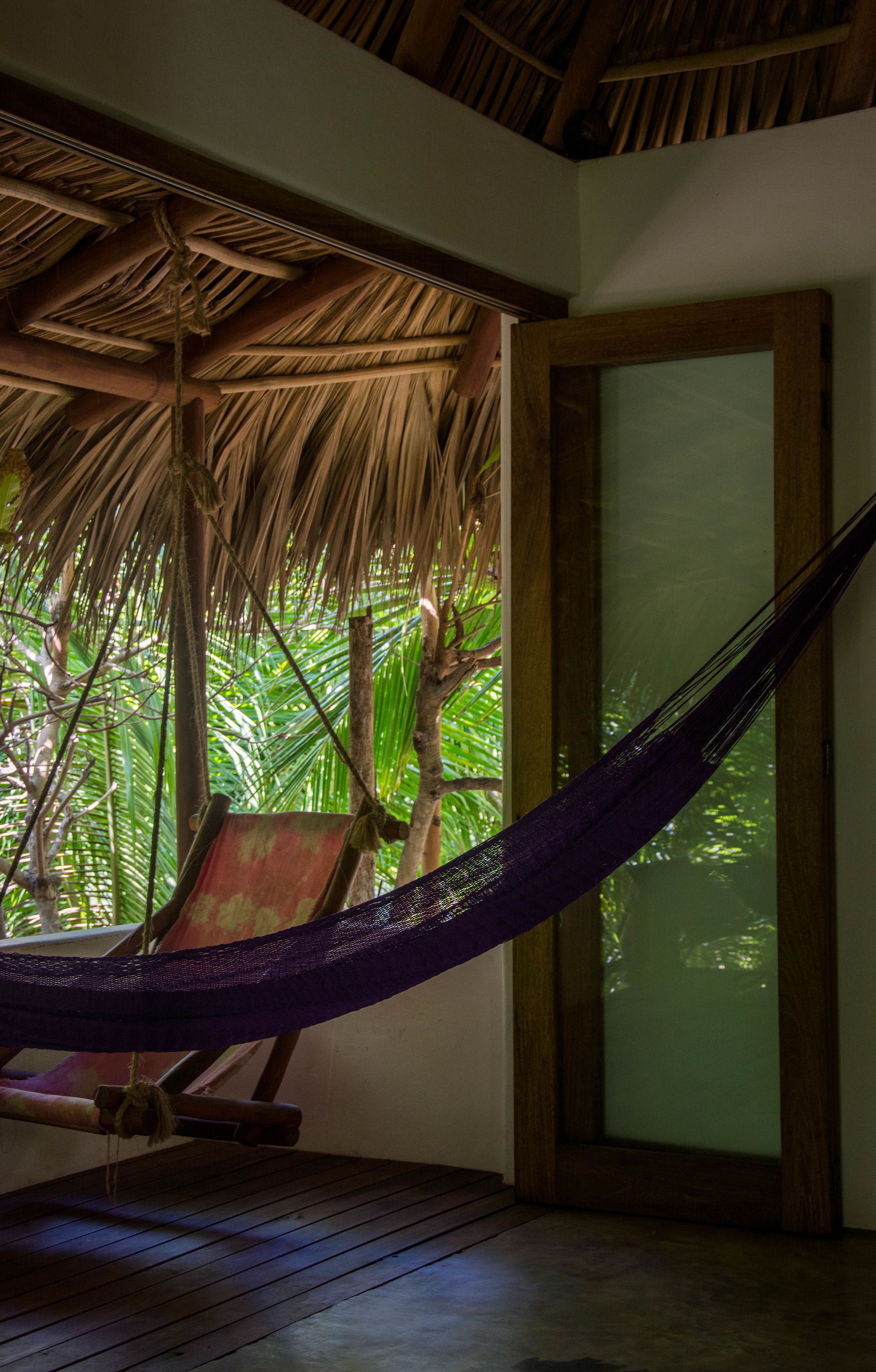 A hammock chair under a thatched roof, wooden flooring, and a view of lush greenery.