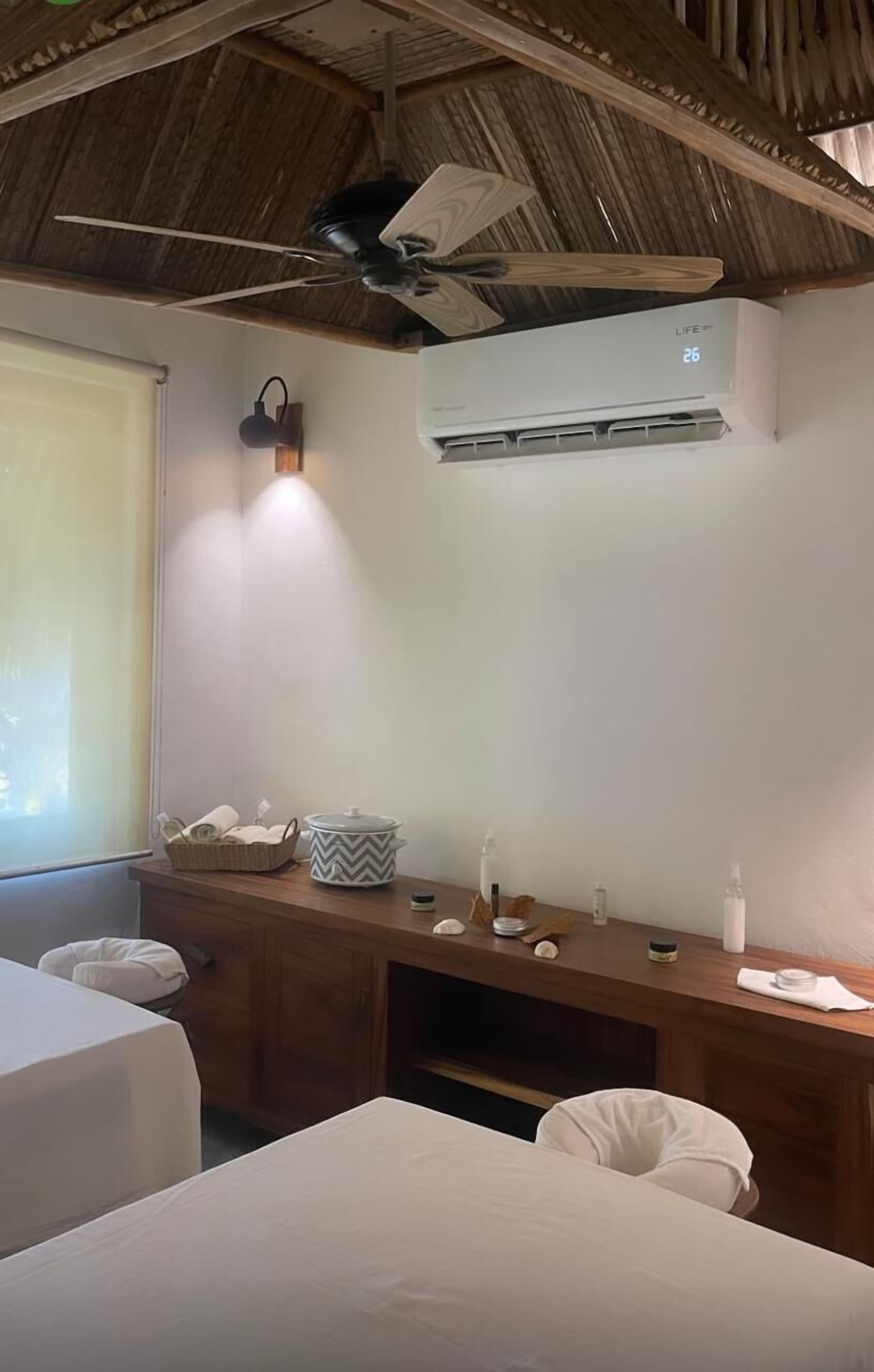 Habitación de hotel con dos camas, un escritorio de madera con artículos de aseo, un ventilador de techo y un equipo de aire acondicionado.