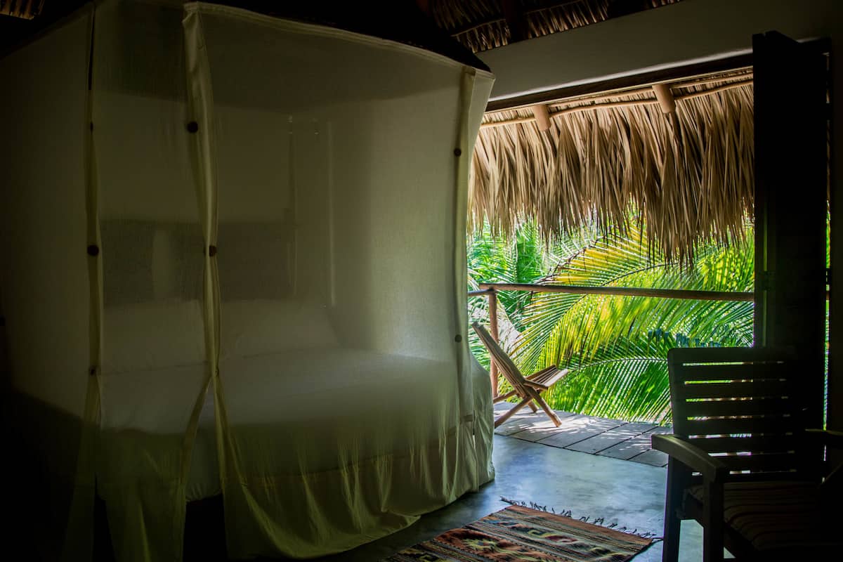 Ein Zimmer mit einem Moskitonetz über dem Bett, einem Holzstuhl und Blick auf eine Palme durch ein Reetdach.