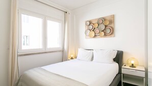 A neatly arranged bedroom with a large bed, two bedside tables with lamps, a window with curtains, and a wall-mounted decorative piece.