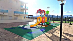 Un parque infantil con tobogán y estructuras para escalar, rodeado de una zona pavimentada con vistas a un edificio y montañas al fondo.