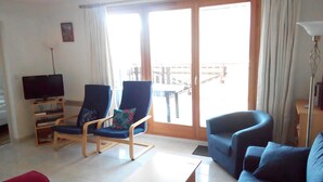 Een woonkamer met een televisie, twee fauteuils, een bank en een balkon met een leuning.