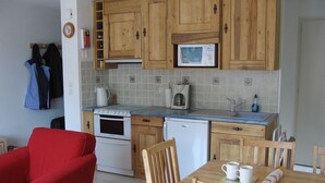 Een kleine keuken met houten kastjes, een fornuis, een magnetron en een spoelbak. In de woonkamer staat een eettafel met stoelen en een rode bank.