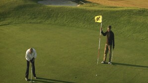 Ein Golfplatz mit Fahnenstange, einem Sandbunker und Blick auf Strand und Meer im Hintergrund.
