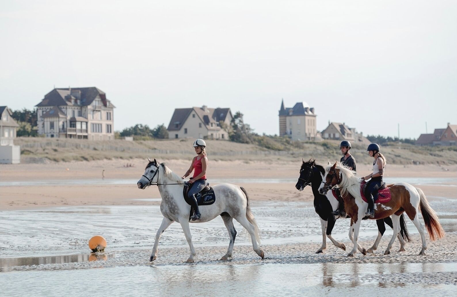 Drei Personen reiten Pferde am Strand, im Hintergrund Häuser.