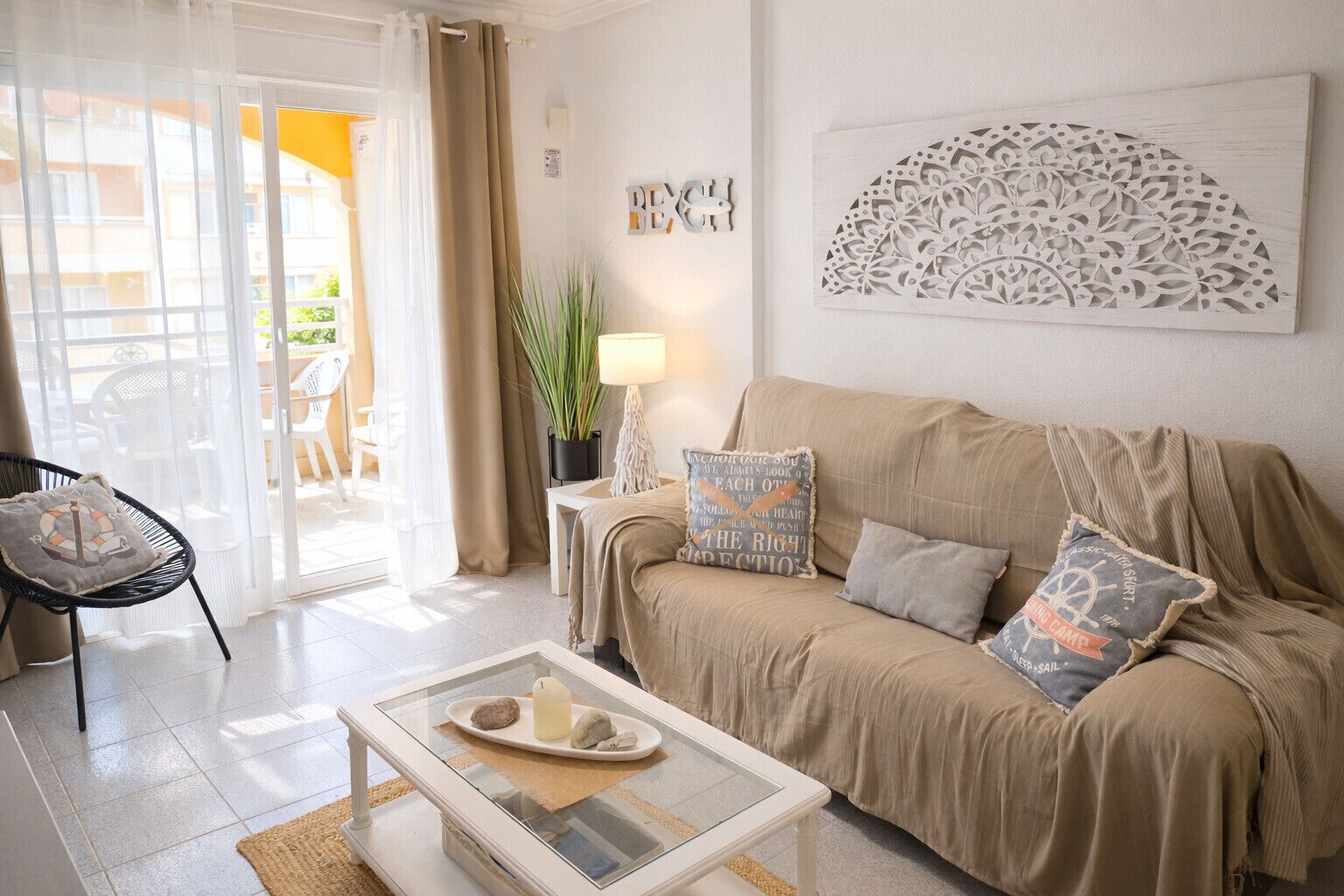 Una sala de estar con un sofá beige, una mesa de centro de cristal con objetos decorativos, una silla y vistas a través de un ventanal.