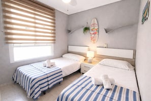 Une chambre d’hôtel avec deux lits, une fenêtre avec des stores, une décoration sur le thème du surf et une table de chevet avec une lampe.