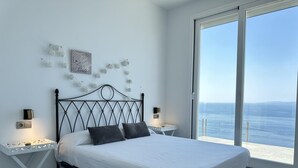 Une chambre à coucher comprenant un lit, deux tables de chevet, une lampe fixée au mur et une vue sur la mer par une grande fenêtre.