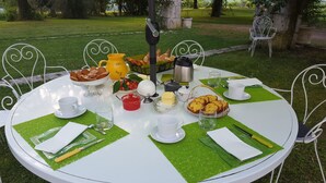 A round table set outdoors with a variety of food items, including bread, butter, and a pitcher, surrounded by white chairs.