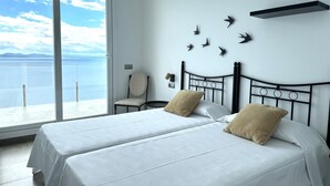Une chambre avec deux lits, une chaise, une étagère fixée au mur et une vue sur la mer.