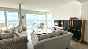 Un salon moderne doté d’un grand téléviseur, de canapés à rayures et offrant une vue sur la mer grâce à de grandes fenêtres.
