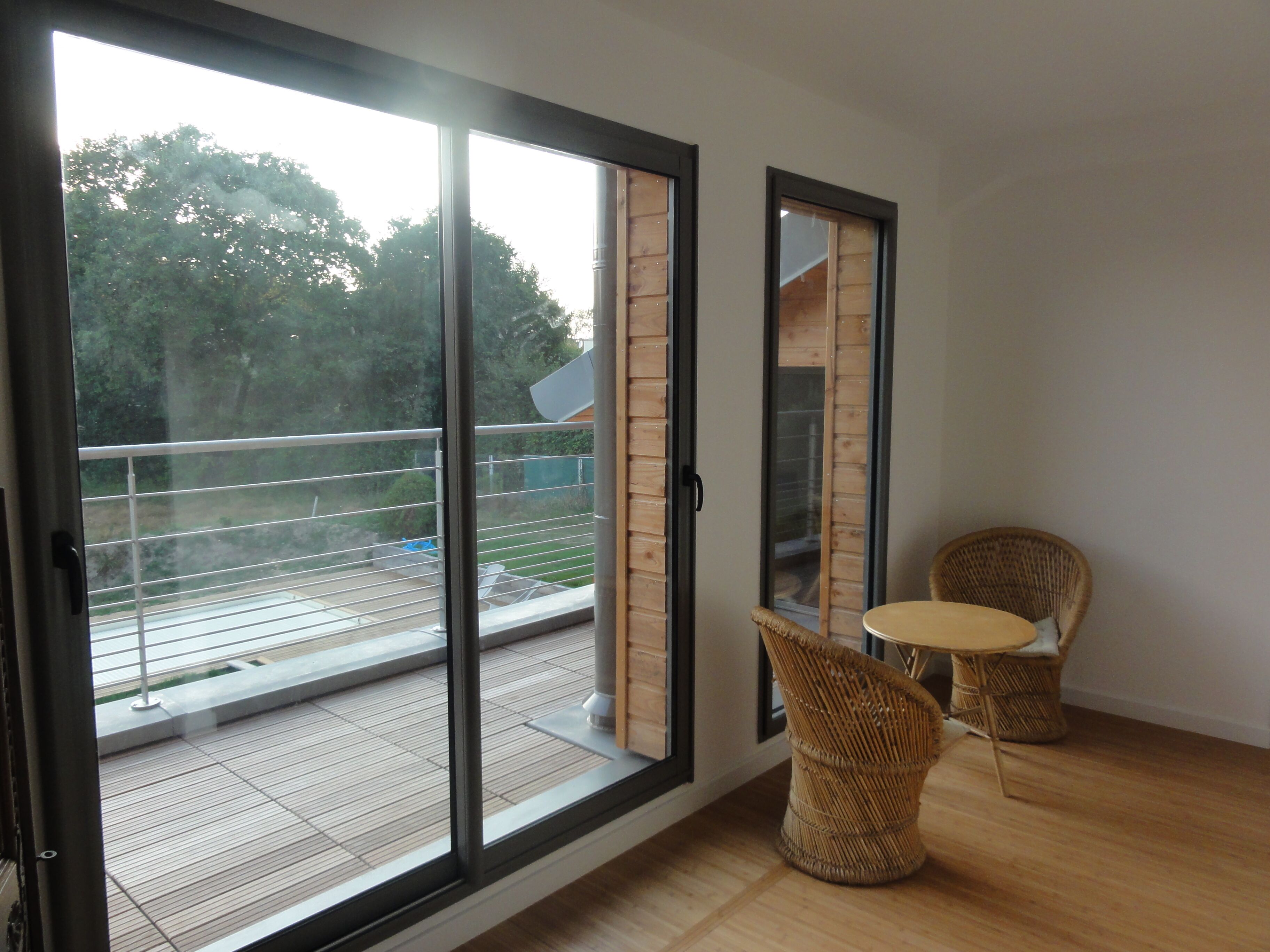Ein Zimmer mit einer Schiebetür, die auf einen Außenbalkon mit Blick auf Bäume und einen Zaun führt. Auf dem Balkon stehen zwei Korbstühle und ein kleiner runder Tisch.