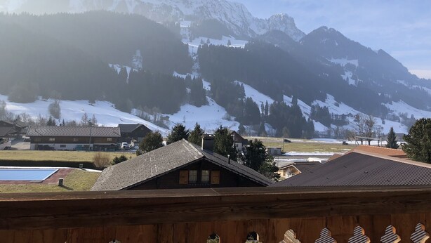 Vista de la montaña desde una cabaña de madera, con picos nevados y un pequeño pueblo en la falda.