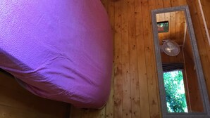 Una habitación con suelo de madera, un objeto tapizado en tela morada, un ventilador de techo y una ventana con vista a la vegetación.