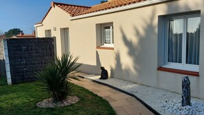 Una casa con techo de tejas, un pequeño jardín con una planta de palmera y una estatua de una persona sentada en el suelo.
