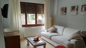 Een woonkamer met een witte bank, rode poefs, een kleine tafel met een decoratief object, een aan de muur bevestigde televisie en een raam met zonwering.