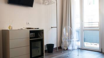 Ein Zimmer mit einem flachbildfernseher an der Wand, einer Holzkabine mit Schubladen, einem klaren Kunststoffstuhl, einer schwarzen Mülltonne und einem großen Fenster mit Ausblick.