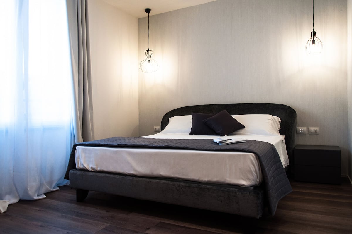 Un dormitorio moderno con una cama grande, dos lámparas colgantes, una mesita de noche y una ventana con cortinas.
