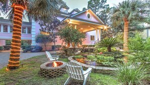 A house with a fire pit and outdoor seating, surrounded by a garden with a pond and palm trees.