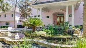A pink building with a covered porch, a small pond, and a palm tree.