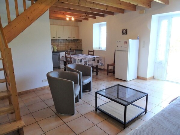 A living room with a glass coffee table, two grey armchairs, a wooden staircase, a kitchen in the background, and a white refrigerator.