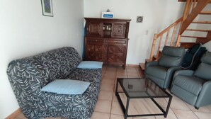 Una sala de estar con un sofá estampado, una mesa de centro de cristal y un sofá verde. Un mueble de madera con un letrero está apoyado en la pared.